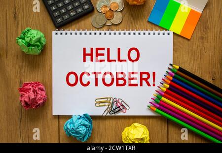 Note blanche avec inscription « bonjour octobre » sur une belle table en bois, papier coloré, crayons de couleur, trombones, pièces de monnaie et calculatrice. Business con Banque D'Images