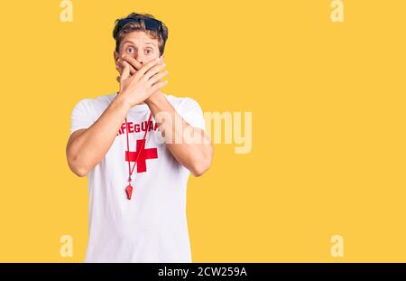 Jeune homme beau portant le t-shirt de maître-nageur et sifflet choqué couvrant la bouche avec des mains pour erreur. Concept secret. Banque D'Images