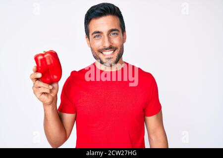 Jeune hispanique tenant le poivron rouge, il a l'air positif et heureux debout et souriant avec un sourire confiant montrant les dents Banque D'Images