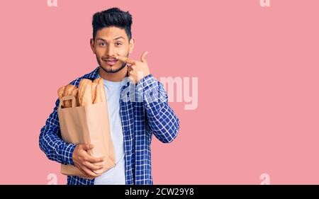 Beau jeune homme latino-américain tenant un sac de papier avec du pain pointant du doigt vers le visage et le nez, souriant gai. Concept de beauté Banque D'Images