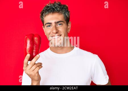 Jeune homme beau tenant le poivron rouge, il a l'air positif et heureux debout et souriant avec un sourire confiant montrant les dents Banque D'Images