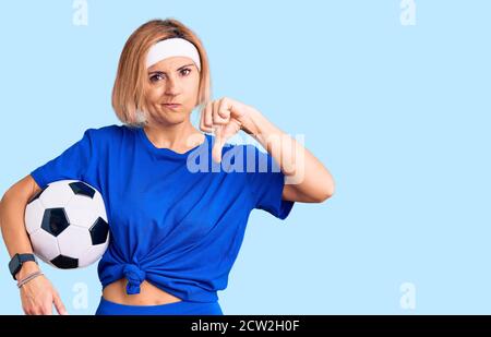 Jeune femme blonde tenant le ballon de football avec le visage en colère, signe négatif montrant le désarroi avec les pouces vers le bas, concept de rejet Banque D'Images