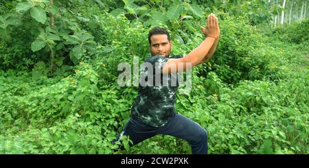 DISTRICT KATNI, INDE - 02 SEPTEMBRE 2019 : un garçon indien faisant du yoga virabhadrasana pose sur fond de forêt de feuilles vertes. Banque D'Images