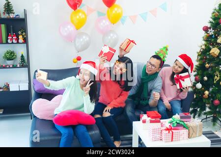 Groupe de collègues multiethniques prenant le selfie dans les chapeaux de Santa lors de la fête de bureau, les jeunes dames célèbrent la fête dans la maison pleine de ballons colorés et de prese Banque D'Images