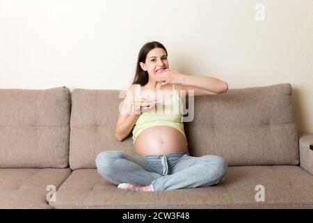 La femme enceinte est en train de manger beaucoup de beignes se relaxant sur le canapé. Diète malsaine pendant la grossesse concept. Banque D'Images