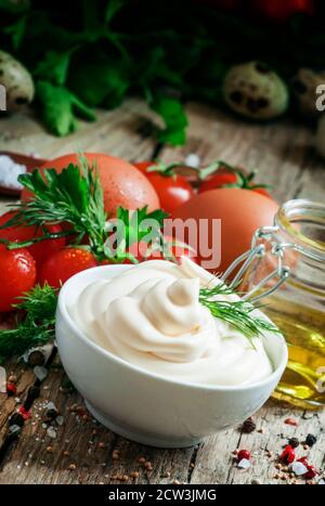 Sauce mayonnaise maison dans un bol blanc, bol avec huile d'olive, œufs, sel, épices, moutarde, herbes, tomates cerises sur fond de bois ancien, sélective Banque D'Images