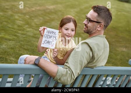 Célébrez la fête des Pères heureux en plein air. Jeune père aimant assis sur le banc en bois dans le parc avec sa petite fille mignonne, heureuse fille tenant Banque D'Images