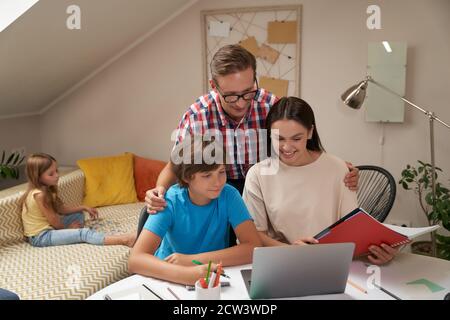 Homeschooling. De jeunes parents positifs aidant son fils à faire ses devoirs tandis que sa petite sœur est assise sur un canapé sur le fond et jouant avec des puzzles Banque D'Images