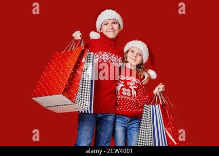 Enfants adorables en chapeaux de père Noël rouges, chandails tenant des sacs de shopping sur fond rouge. Banque D'Images