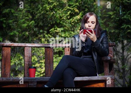 Jeune femme dans une veste en cuir assise sur un banc et jouant sur un smartphone dans le parc. La brunette adulte repose parmi les arbres verts dans le parc de la ville et la navigation Banque D'Images