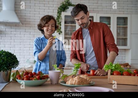 Passer un week-end à la maison. Un jeune papa et un petit fils heureux coupent des légumes frais, préparant une salade ensemble tout en se tenant dans la cuisine moderne Banque D'Images