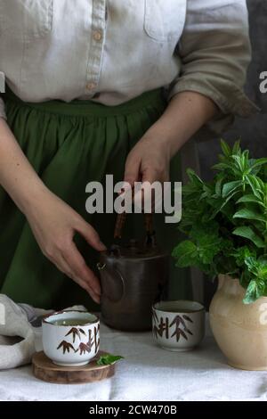 Thé à la menthe dans un ensemble de thé chinois vintage, une femme dans une robe de lin et un tablier, et un bouquet de menthe dans une carafe. Banque D'Images