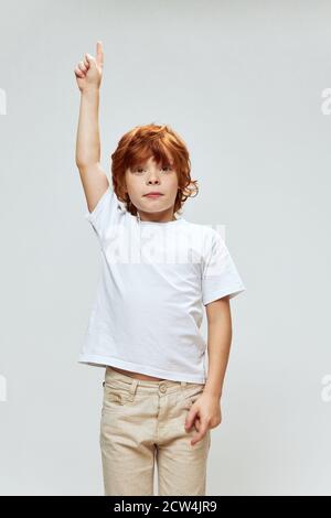 l'enfant à cheveux rouges tient sa main au-dessus de sa tête pointant vers L'espace de copie de l'arrière-plan gris supérieur Banque D'Images