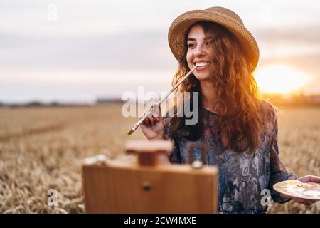 Belle fille artiste peint dans un champ de blé. Une femme sourit, tient une brosse et une palette dans les mains, devant son chevalet. Derrière elle est orange Banque D'Images