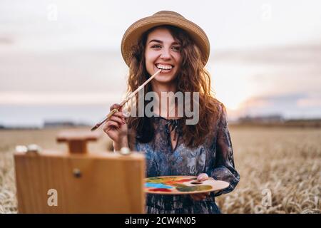 Belle fille artiste peint dans un champ de blé. Une femme sourit, tient une brosse et une palette dans les mains, devant son chevalet. Derrière elle est orange Banque D'Images