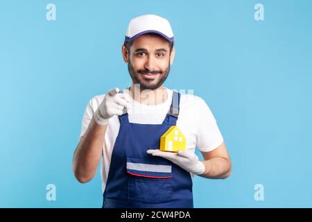 Hé ! Portrait d'un homme de main heureux, un homme de mouvement en vêtements de travail et des gants tenant la maison de papier et pointant vers l'appareil photo. Publicité réparation de maison, entretien Banque D'Images