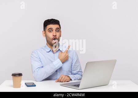 Regardez, faites de la publicité ici! Stupéfait homme d'affaires choqué assis bureau avec ordinateur portable sur bureau, pointant de côté espace de copie vierge pour l'idée commerciale Banque D'Images