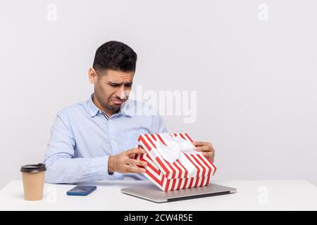 Un employé malheureux assis dans un bureau, ouvrant une boîte cadeau et regardant à l'intérieur avec une expression triste déçue, déballant le mauvais présent, celebr Banque D'Images