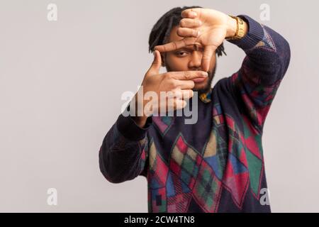 Homme africain sérieux avec des dreadlocks regardant à travers les doigts faisant des gestes d'appareil photo, les hommes d'appareil photo à la recherche d'une mise au point correcte, capturant des moments. St. Intérieure Banque D'Images