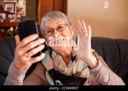 Femme sénior souriante à la maison agitant comme elle fait la vidéo Appelez votre famille sur votre téléphone portable Banque D'Images