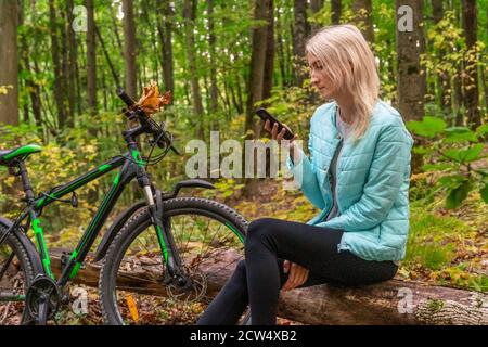 Blonde fille dans une veste turquoise est assis sur un arbre tombé dans la forêt d'automne et regarde le téléphone et sourit, il y a un vélo à côté d'elle Banque D'Images