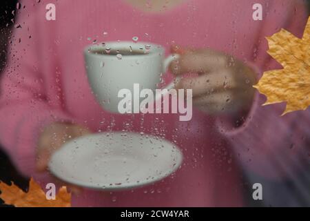 tasse blanche dans la main et la fenêtre avec gouttes de pluie.arrière-plan flou. mise au point sélective. Banque D'Images