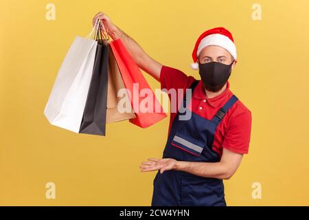 Le livreur dans le chapeau du père noël et masque de protection noir montrant les sacs d'achats, la commande facile de marchandises et la livraison en quarantaine. Prise de vue en studio I Banque D'Images