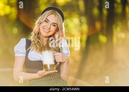 Jolie blonde heureuse dans dirndl, robe traditionnelle de festival, tenant une tasse de bière à l'extérieur dans la forêt avec un fond flou. Oktoberfest, Saint-Patric Banque D'Images