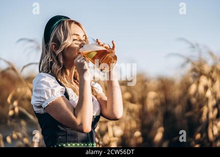 Jolie blonde heureuse dans dirndl, robe traditionnelle de festival, boire de la bière à l'extérieur dans le champ avec un fond flou. Oktoberfest, jour de la Saint-Patrick Banque D'Images