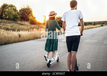 Couple heureux marchant sur des scooters. Une jeune femme en robe et chapeau sur fond de coucher de soleil. Au premier plan, un homme tourne son dos sur l'objectif. Banque D'Images