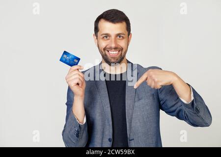 Jeune homme d'affaires avec barbe en veste tenant une carte de crédit debout sur fond blanc isolé avec le visage surpris pointant du doigt vers lui. Portrait d'un homme sur fond gris. Banque D'Images