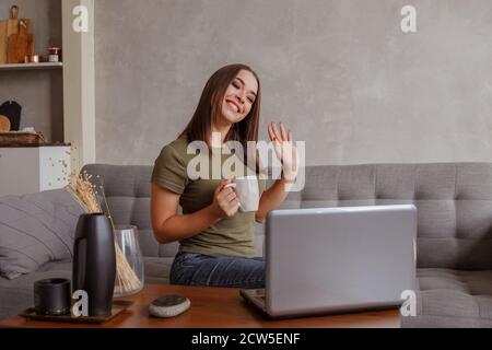 Portrait d'une jeune femme agréable avec une tasse regardant l'appareil photo, en agitant Bonjour. Jolie femme du millénaire communiquant avec ses parents par vidéoconférence Banque D'Images