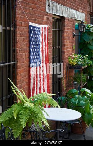 BROOKLYN, NY - SEPTEMBRE 23 2020 : un drapeau américain est suspendu sur le côté d'une ancienne maison en brique, dans un petit jardin entre les plantes et une table et des chaises en fil métallique Banque D'Images