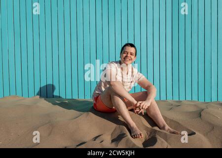 Jeune homme assis sur une plage de sable souriant à l'appareil photo un mur turquoise Banque D'Images