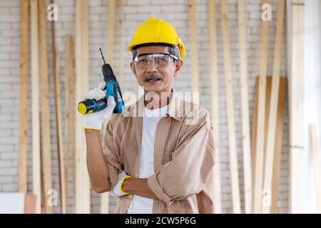 Homme asiatique senior avec casque de sécurité et lunettes de sécurité tient la perceuse électrique dans l'atelier de menuiserie. Concept senior personnes faisant hobby après la retraite. Banque D'Images