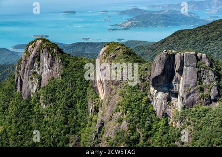 Vue rapprochée et détail des trois pics de formation rocheuse couverts de la végétation verte de Serra do Mar avec la côte de la baie de Carioca en arrière-plan lointain. Banque D'Images