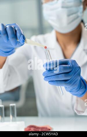 Gloved chercheur en whitecoat et masque mettant un petit morceau de viande dans un flacon Banque D'Images