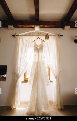 Longue robe blanche de la mariée sur un cintre sur le cornice avec des chaussures pour femmes à la fenêtre avec des rideaux ouverts dans la chambre. Banque D'Images