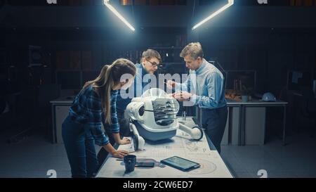 Tard dans la nuit dans l'installation d'ingénierie robotique trois ingénieurs techniques parlent et travaillent sur un prototype de robot à roues. En arrière-plan High Tech Banque D'Images