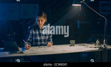 Une femme ingénieure assise à son bureau travaille avec des Blueprints sur une table, utilise un crayon, une règle et une tablette numérique. Dans la conception industrielle sombre Banque D'Images