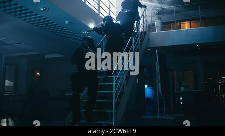 Une équipe masquée d'officiers de police SWAT armés descend les escaliers depuis un deuxième étage dans un immeuble de bureaux sombre. Soldats avec des fusils et des lampes de poche Banque D'Images