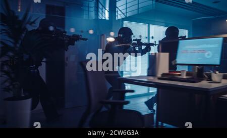 Une équipe masquée d'officiers de police SWAT armés se déplace dans une salle d'un immeuble de bureaux saisi par les ténèbres avec des bureaux et des ordinateurs. Soldats avec fusils et Banque D'Images