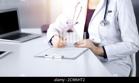 Une femme-médecin inconnue prescrit des médicaments à son patient, à l'aide d'un presse-papiers, tout en étant assise au bureau dans son cabinet ensoleillé. Femme Banque D'Images