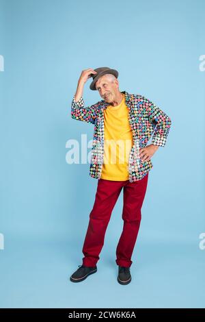 Salutation joyeuse. Portrait de l'homme hipster senior dans un chapeau élégant isolé sur fond bleu studio. Concept de mode de vie technologique et joyeux pour les personnes âgées. Couleurs tendance, toujours jeune. CopySpace pour publicité. Banque D'Images