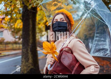 Femme élégante en masque de protection marche dans la rue d'automne ville sous un parapluie transparent pendant la pluie. Pandémie de Covid Banque D'Images