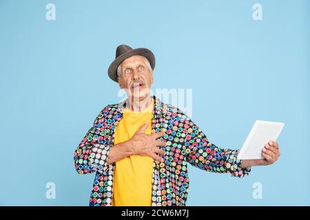 Chat vidéo avec tablette, choqué. Portrait de l'homme hipster senior dans un chapeau élégant sur fond bleu studio. Concept de mode de vie technologique et joyeux pour les personnes âgées. Couleurs tendance, toujours jeune. CopySpace pour publicité. Banque D'Images