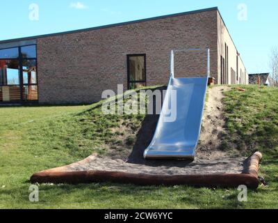 Un toboggan en métal près du centre après l'école et de la maternelle. L'herbe est verte, le ciel est bleu et un bâtiment en brique rouge est visible en arrière-plan. Banque D'Images