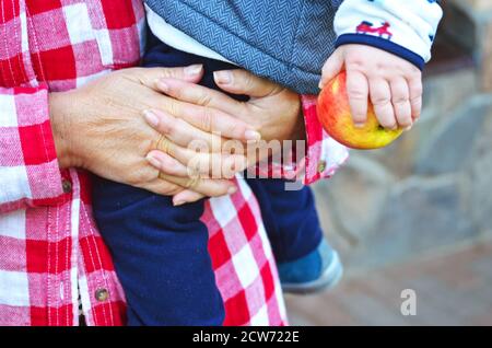 Gros plan. Les mains de la grand-mère se sont froissées, tenant son petit-fils. Bébé tient une pomme. Vêtements chauds, chemise à carreaux rouges. Le temps de la famille, les générations Banque D'Images