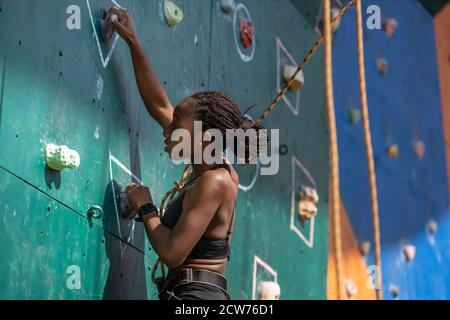 Jeune femme noire grimpant sur le mur d'escalade d'entraînement. Fille grimpeuse africaine qui traverse la route Banque D'Images