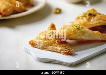 Dessert turc traditionnel maison, baklava avec pistache, servi sur une planche à découper en bois blanc sur une table en marbre blanc. Banque D'Images